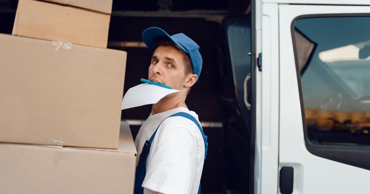 Ένας επαγγελματίας φορτώνει χάρτινες κούτες σε φορτηγάκι. Με το στόμα του κρατάει χαρτιά και στυλό.