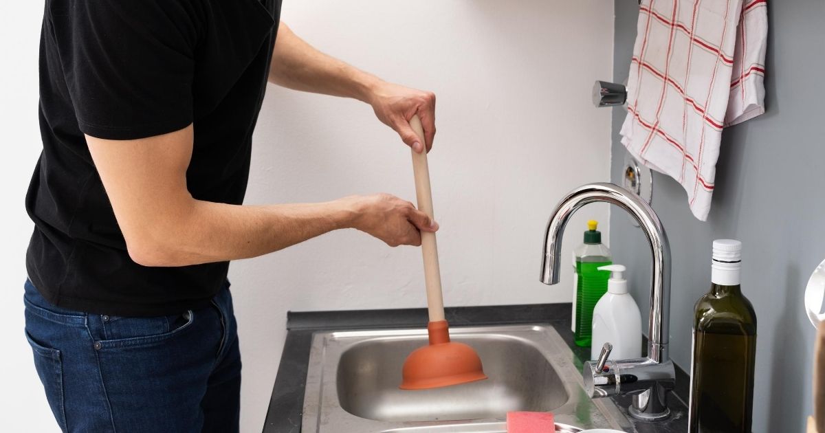 Ένας άνδρας μπροστά από νεροχύτη κουζίνας, κρατάει μια βεντούζα ξεβουλώματος.