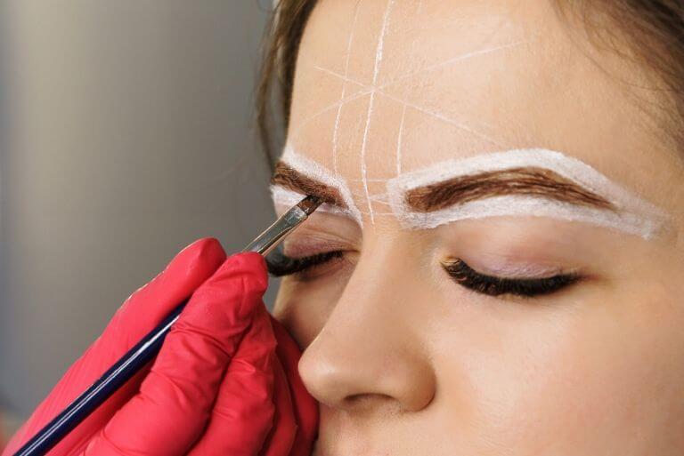 Επαγγελματίας φορώντας κόκκινο γάντι, βάφει με πινελάκι τα φρύδια σε γυναικείο πρόσωπο.