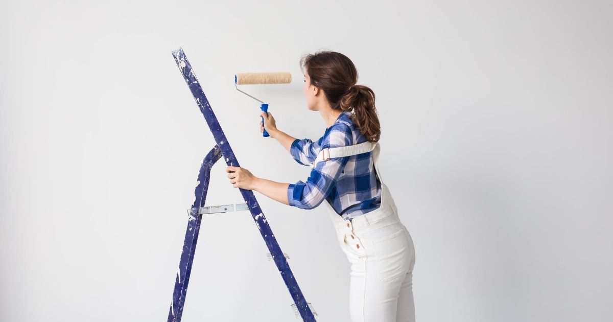 Μια γυναίκα επάνω σε σκάλα, βαφει τον τοίχο με ρολό.